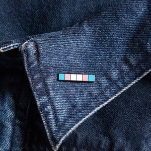 Pode incluir: Uma jaqueta de ganga azul com um broche de orgulho transgénero no colarinho. O broche é uma barra horizontal com listras azul claro, rosa e azul claro.