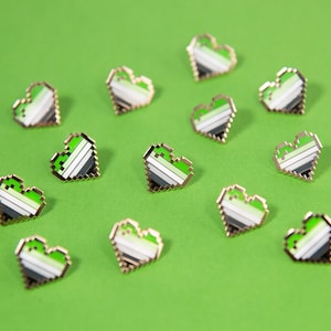 May include: A collection of 12 green, white, and black pixelated heart pins. The pins are arranged in a scattered pattern on a green background.