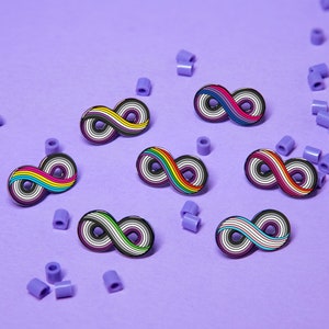 Puede incluir: Varios pines con el símbolo del infinito, cada uno con diseños de banderas del orgullo LGBTQ+ (arcoíris, bisexual, asexual). Los pines están sobre un fondo morado con cuentas moradas dispersas.