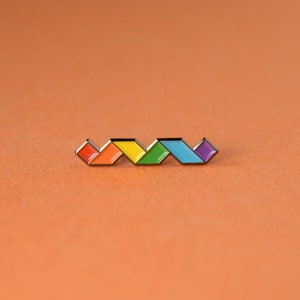 May include: A colorful enamel pin featuring a zig-zag design of six connected diamond shapes in a rainbow of colors: red, orange, yellow, green, blue, and purple. The pin is set against an orange background.