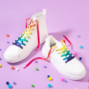 May include: A pair of white sneakers with rainbow-colored laces. One is a high-top, the other a low-top. The laces are a gradient of colors. Colorful pom-poms are scattered on the purple surface.