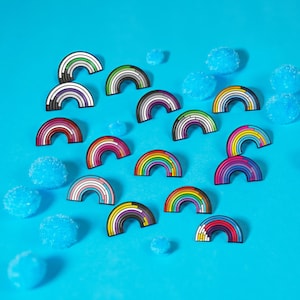 May include: A collection of rainbow-shaped enamel pins with various color schemes, including pride flag designs, scattered on a bright blue surface. The pins are black-outlined and feature different rainbow patterns. Blue, spherical decorations are also present.