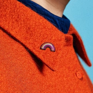 May include: An orange jacket with a small rainbow pin on the collar. The jacket has a pocket and buttons down the front. A blue shirt is visible underneath the jacket. The background is a solid light blue.
