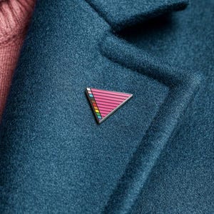 May include: A teal wool coat with a pink turtleneck sweater underneath. A silver-toned triangle pin with pink and rainbow stripes is pinned to the coat. The background is a solid light blue.