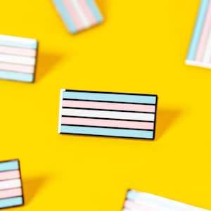 May include: Rectangular enamel pins with the transgender pride flag design. The pins feature horizontal stripes in light blue, pink, and white, outlined in black. The pins are scattered on a bright yellow background.