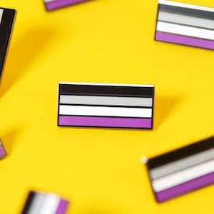 May include: Multiple black, white, and purple asexual pride flag pins on a yellow background.
