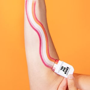 以下が含まれることがあります： 虹色のデザインが描かれた腕が、"THE PIN PEOPLE"の文字が入った白い長方形のオブジェクトで保持されています。その人はもう一方の手で平和のサインをしています。背景はオレンジ色です。縞模様のトップスを着ています。