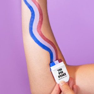 May include: An arm with red, white, and blue paint strokes, like a flag, applied from a small white applicator labelled "THE PIN PRICE". The person is making a rock-on hand gesture against a purple background.