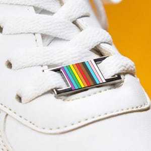 May include: Close-up of a white sneaker with white laces and a colorful rainbow-striped accessory on the laces. The shoe is set against a bright yellow background, highlighting the details of the footwear.
