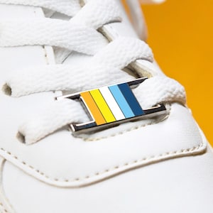 May include: A close-up shot of a white trainer with white laces and a decorative shoe tag. The tag has vertical stripes in yellow, orange, white, and blue. The background is a bright yellow.