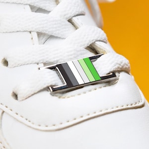 May include: A close-up shot of a white trainer with white laces and a decorative metal tag. The tag has a design with black, grey, white, and green stripes. The shoe is set against a bright yellow background.