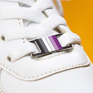 May include: A close-up of a white trainer with white laces and a silver lace tag. The lace tag displays the asexual pride flag colours: black, grey, white, and purple. The background is a solid yellow.