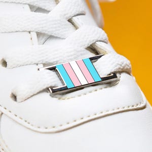 Op de afbeelding: Close-up van een witte sneaker met witte veters en een decoratieve metalen staaf op de veters. De staaf heeft een transgender pride vlag ontwerp in lichtblauw, roze en wit. De achtergrond is effen geel.
