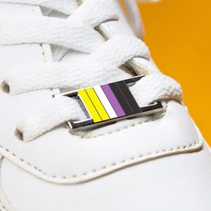 Op de afbeelding: Close-up van een witte sneaker met witte veters en een non-binaire pride flag veter tag. De tag heeft gele, witte, paarse en zwarte strepen. De schoen staat tegen een felgele achtergrond.