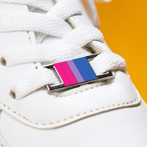 May include: A close-up shot of a white trainer with white laces and a decorative metal tag. The tag has a horizontal stripe design in pink, purple, and blue. The background is bright yellow. The shoe appears clean and new.