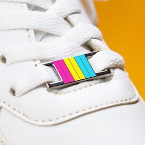 May include: A white trainer with white laces and a decorative shoe tag. The tag has pink, yellow, and blue stripes. The image is set against a bright yellow background.