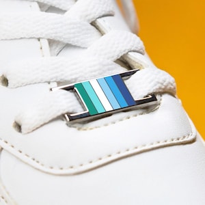 Puede incluir: Primer plano de una zapatilla blanca con cordones blancos y una etiqueta metálica decorativa. La etiqueta presenta rayas horizontales en tonos verdes y azules. El fondo es amarillo liso.