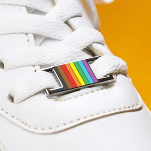 May include: A close-up shot of a white trainer with white laces and a rainbow-coloured shoe tag. The tag is rectangular with a silver border and features a vertical stripe design in rainbow colours. The shoe is set against a yellow background.