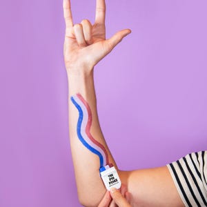 May include: A person's arm with red, white, and blue paint strokes, resembling a flag, applied from a small white applicator labeled "THE PIN PRICE". The person is making a rock-on hand gesture against a purple background.