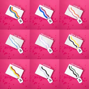 May include: Nine white cards with colorful paint strokes representing different identities, including rainbow, bisexual, lesbian, gay man, transgender, non-binary, pansexual, asexual, and aromantic. Each card has a paint tube with the text "The Pride Press". The background is pink.