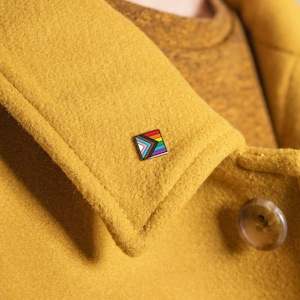 May include: A square enamel pin featuring the Progress Pride flag design. The pin is attached to a mustard-yellow coat. The pin's design includes the traditional rainbow colors, along with the colors of the transgender flag and the colors representing marginalized communities of color.