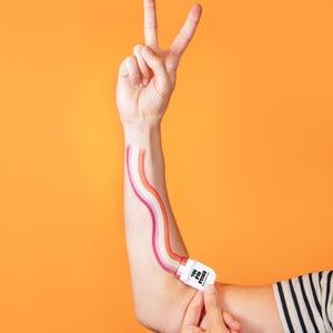 May include: A person's arm with a rainbow-colored design painted on it, held by a white rectangular object with the text "THE PIN PEOPLE". The person is making a peace sign with their other hand. The background is orange.