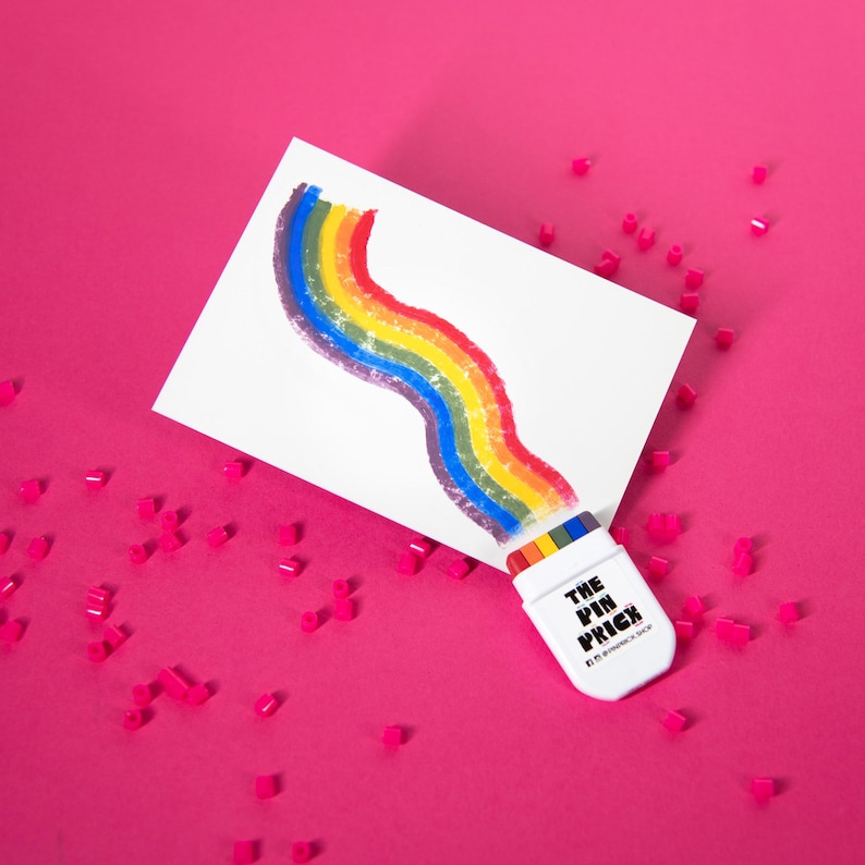 May include: A white card with a rainbow design, with a white container labeled "THE PIN PRICK" at the bottom. The background is a bright pink, scattered with small pink shapes.