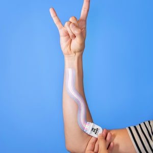 May include: A person's arm with a wavy, pastel-colored design applied with a white applicator. The applicator has the text "THE PIN PATCH" printed on it. The person is making a rock and roll hand gesture against a blue background.