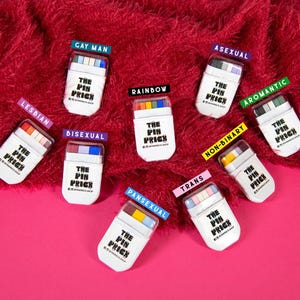 May include: A collection of white rectangular pin cases with colorful tops, each labeled with a different identity, including "LESBIAN," "GAY MAN," "ASEXUAL," and "BISEXUAL." The cases are arranged on a red, textured surface with a pink background. The text "THE PIN PRICK" is printed on each case.