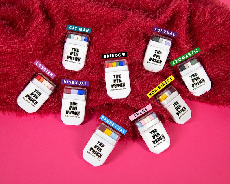 May include: A collection of white rectangular pin cases with colorful tops, each labeled with a different identity, including "LESBIAN," "GAY MAN," "ASEXUAL," and "BISEXUAL." The cases are arranged on a red, textured surface with a pink background. The text "THE PIN PRICK" is printed on each case.