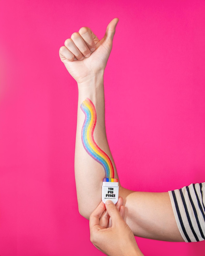 May include: A person's arm with a rainbow painted on it, emerging from a white container held in the hand. The container has the text "THE PIN PRICK" and a rainbow design. The background is a solid pink color.