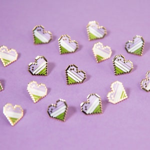 May include: A collection of 12 gold-colored pins shaped like hearts. Each heart is made up of pixelated stripes of lavender, white, and green. The pins are arranged on a purple background.