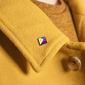 May include: A square enamel pin with a white border, displaying a geometric design in yellow, blue, red, and purple, is attached to a mustard-yellow coat. The pin provides a vibrant contrast against the coat's color.