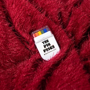 May include: A white rectangular product with a clear lid, containing a rainbow of colors, rests on a textured, deep red surface. The product has the text "THE PIN PRICK" in black and yellow, with social media handles below.