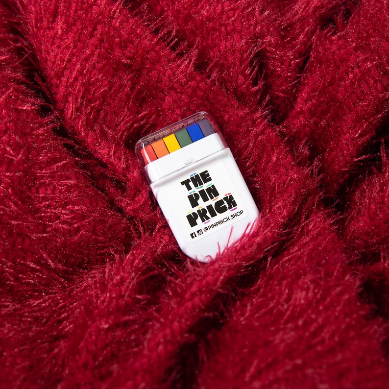 May include: A white rectangular product with a clear lid, containing a rainbow of colors, rests on a textured, deep red surface. The product has the text "THE PIN PRICK" in black and yellow, with social media handles below.