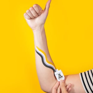 May include: A person's arm with a thumbs-up gesture, featuring a yellow background. The arm has a painted design of black, yellow, and gray wavy lines, connected to a white rectangular pin that reads "THE PIN PRICE".