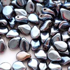 Könnte beinhalten: Nahaufnahme einer großen Menge kleiner, tropfenförmiger Glasperlen. Die Perlen sind dunkel, irisierend grau mit einem metallischen Schimmer.