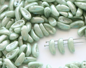 May include: A large quantity of light green glass beads in a teardrop shape. The beads are arranged in a circle around a small group of beads on a white surface.
