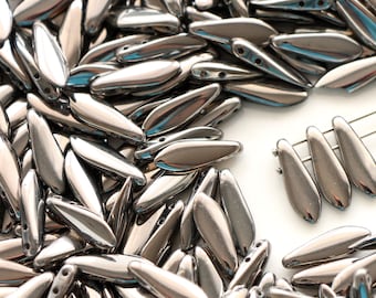 May include: A large pile of silver-colored glass beads in a teardrop shape. The beads have two holes and are scattered on a white surface. A few beads are arranged in a row on the right side of the image.