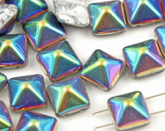 May include: Iridescent square beads with a pyramid design. The beads have a shiny, reflective surface that displays a spectrum of colors, including blues, greens, and yellows. Some beads are strung on silver-colored pins. The beads are on a white background.