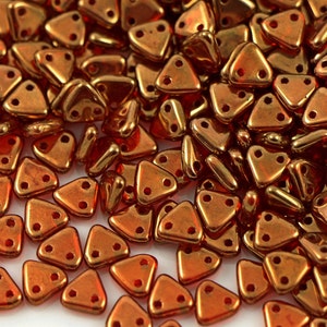 Puede incluir: Primer plano de un montón de pequeñas cuentas de vidrio triangulares de color cobre. Las cuentas tienen dos agujeros cada una y están dispuestas en un patrón aleatorio.