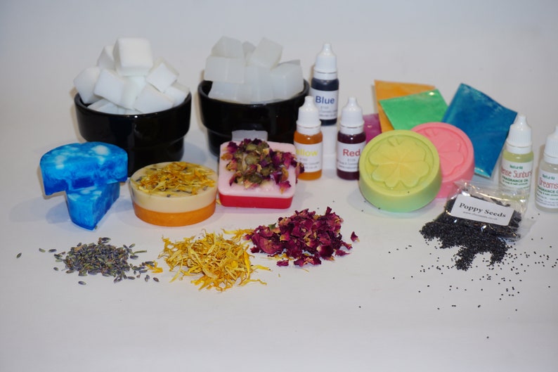 May include: A collection of soap making supplies including white soap base, blue, red, and yellow soap dye, dried rose petals, dried calendula petals, poppy seeds, and a green and yellow soap bar with a clover design.