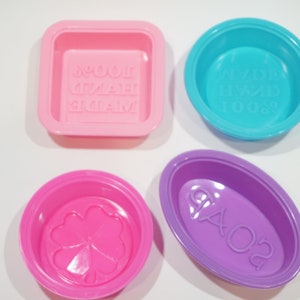 May include: Four silicone soap molds in different shapes and colors. The molds are pink, blue, purple, and pink. The molds have different designs, including a clover, a rectangle with the words "Homemade Soap", a circle with the words "Freshly Made Soap", and an oval with the words "Ciao 02".