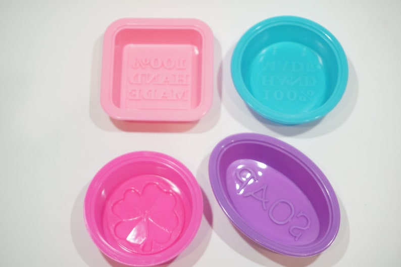 May include: Four silicone soap molds in different shapes and colors. The molds are pink, blue, purple, and pink. The molds have different designs, including a clover, a rectangle with the words "Homemade Soap", a circle with the words "Freshly Made Soap", and an oval with the words "Ciao 02".