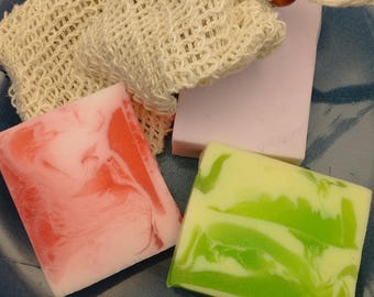 May include: Three bars of soap, one pink and white, one green and white, and one lavender, are displayed on a blue plate. A natural fiber mesh bag with a wooden bead is also shown.