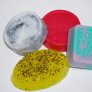 May include: Four handmade soap bars in different colors and shapes. One bar is round and red, one is round and gray with a clover design, one is oval and yellow with black specks, and one is square and green and purple with the text "100% HAND MADE".