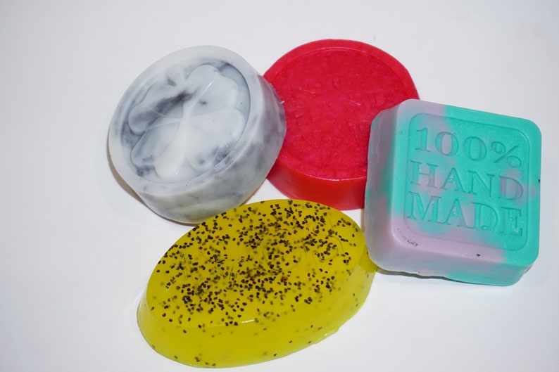 May include: Four handmade soap bars in different colors and shapes. One bar is round and red, one is round and gray with a clover design, one is oval and yellow with black specks, and one is square and green and purple with the text "100% HAND MADE".
