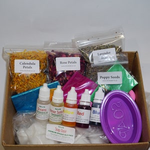 May include: A box filled with ingredients for making soap. The box contains dried flowers, herbs, essential oils, soap base, and a purple soap mold. The box also includes a label that reads "Soap Base Premium Orange SLS Free".