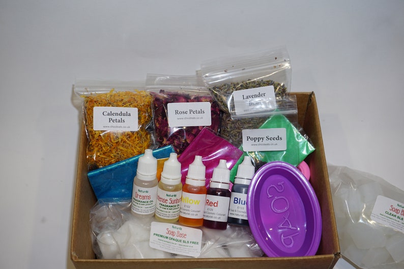 May include: A box filled with ingredients for making soap. The box contains dried flowers, herbs, essential oils, soap base, and a purple soap mold. The box also includes a label that reads "Soap Base Premium Orange SLS Free".