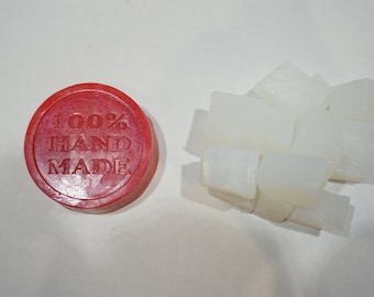 May include: A red round soap base with the text "100% HAND MADE" printed on it. Next to the base are several white soap bars.